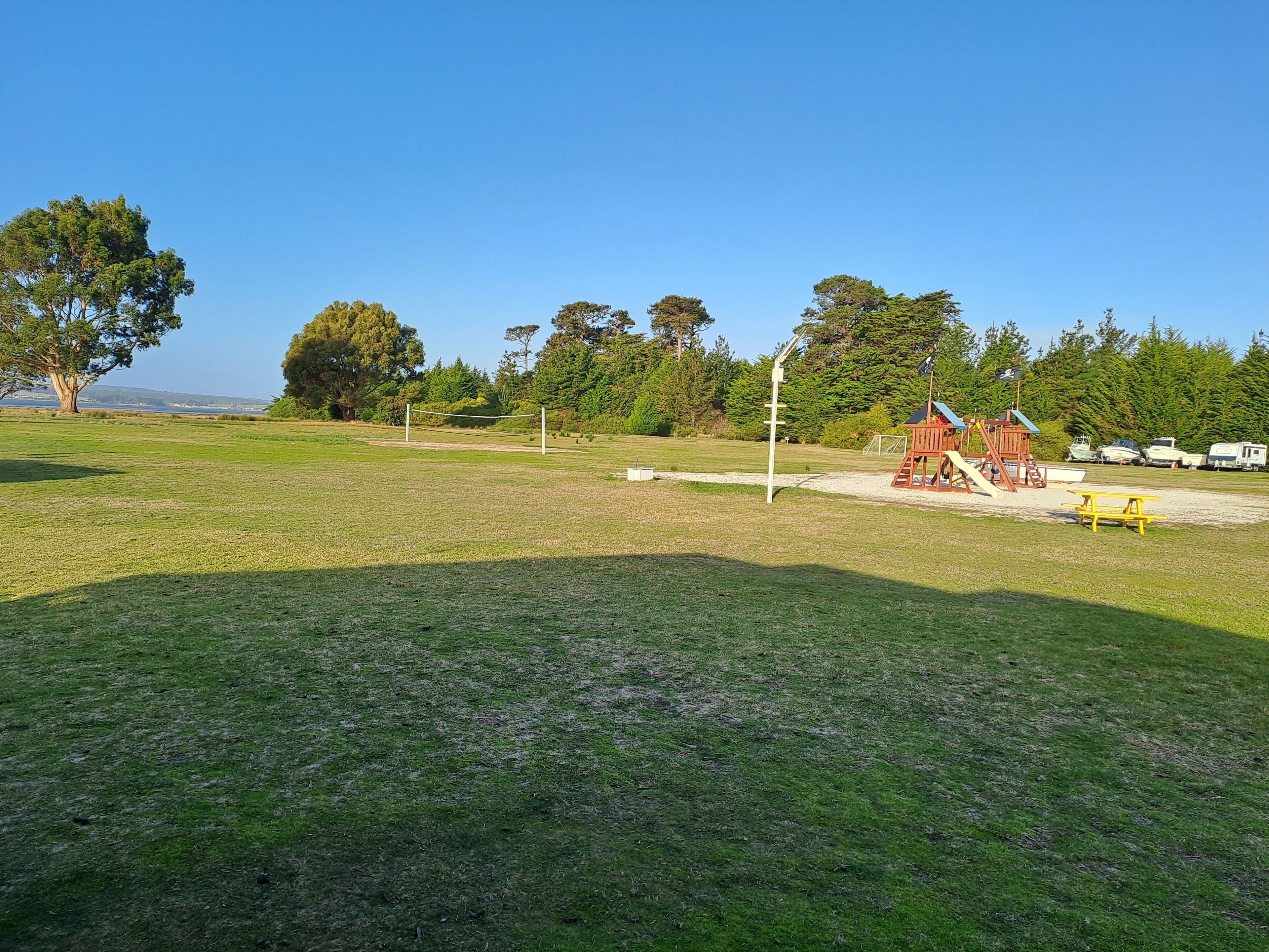 Um parque com gramado, um parquinho, mesa de piquenique e barcos ao fundo.