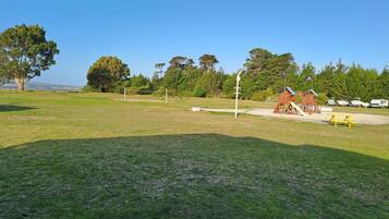 Um parque com gramado, um parquinho, mesa de piquenique e barcos ao fundo.