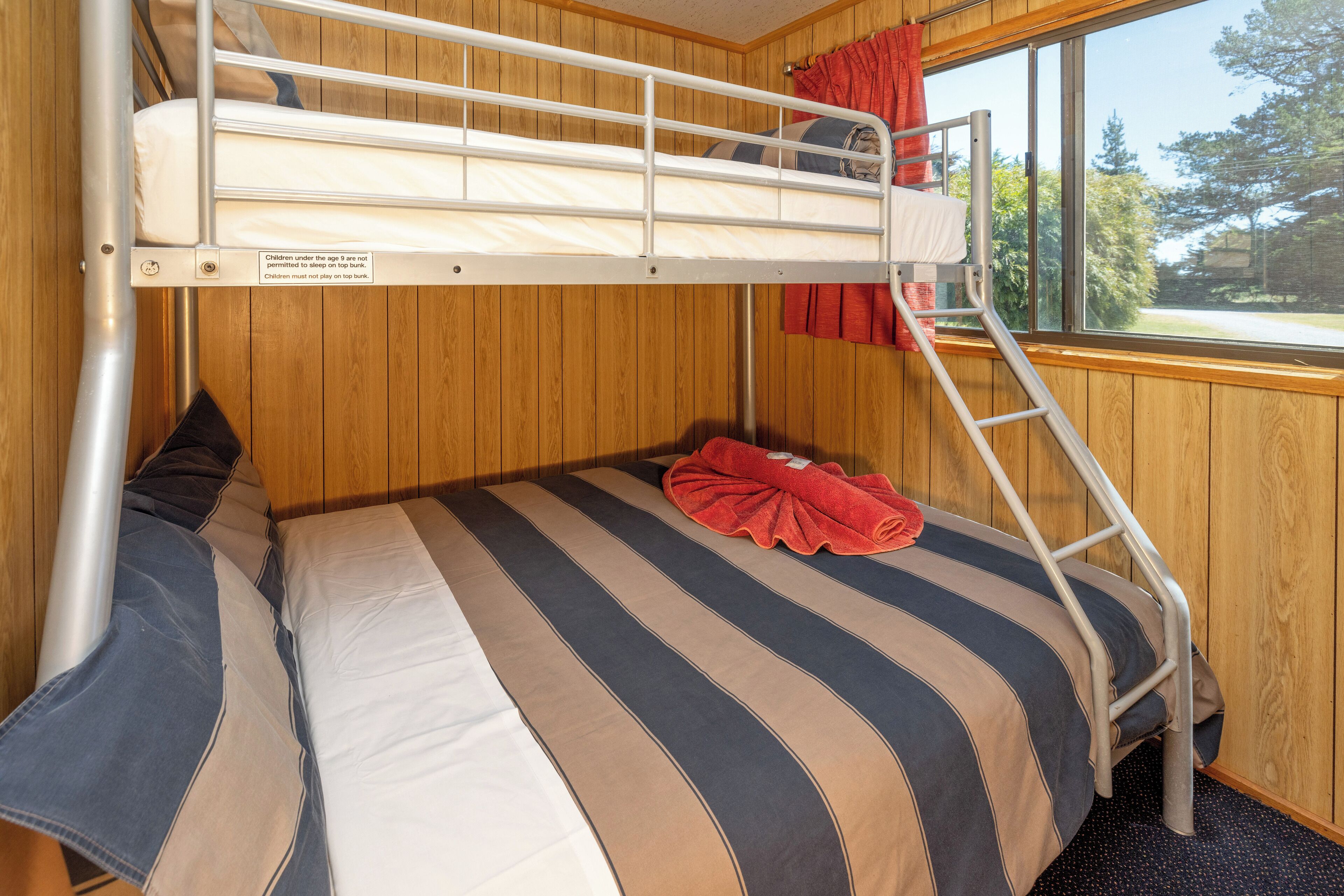 Un lit superposé avec vue sur la fenêtre, des murs en bois et une serviette rouge.