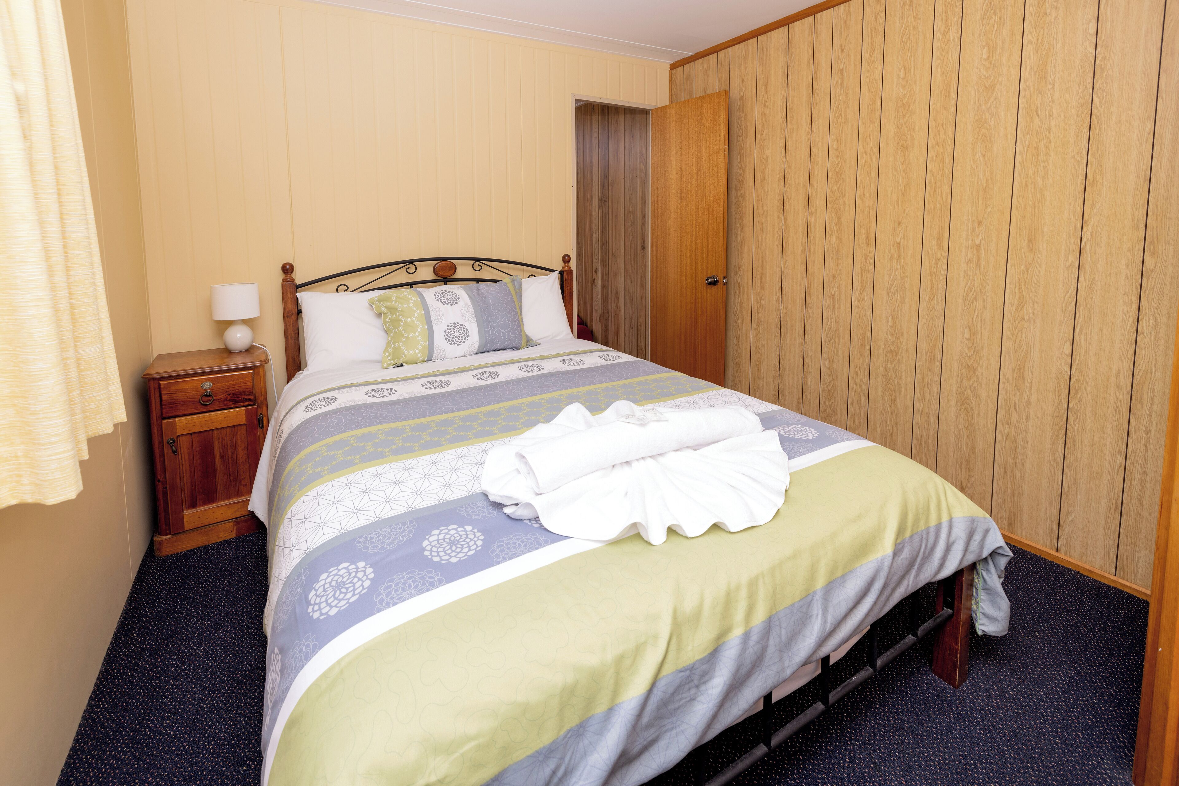 Una cama bien hecha con una colcha estampada, una mesita de noche de madera con una lámpara, y una puerta de madera al fondo.