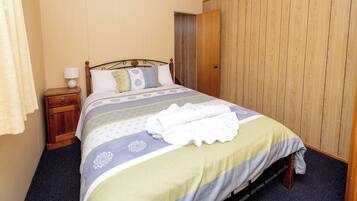 Una cama bien hecha con una colcha estampada, una mesita de noche de madera con una lámpara, y una puerta de madera al fondo.