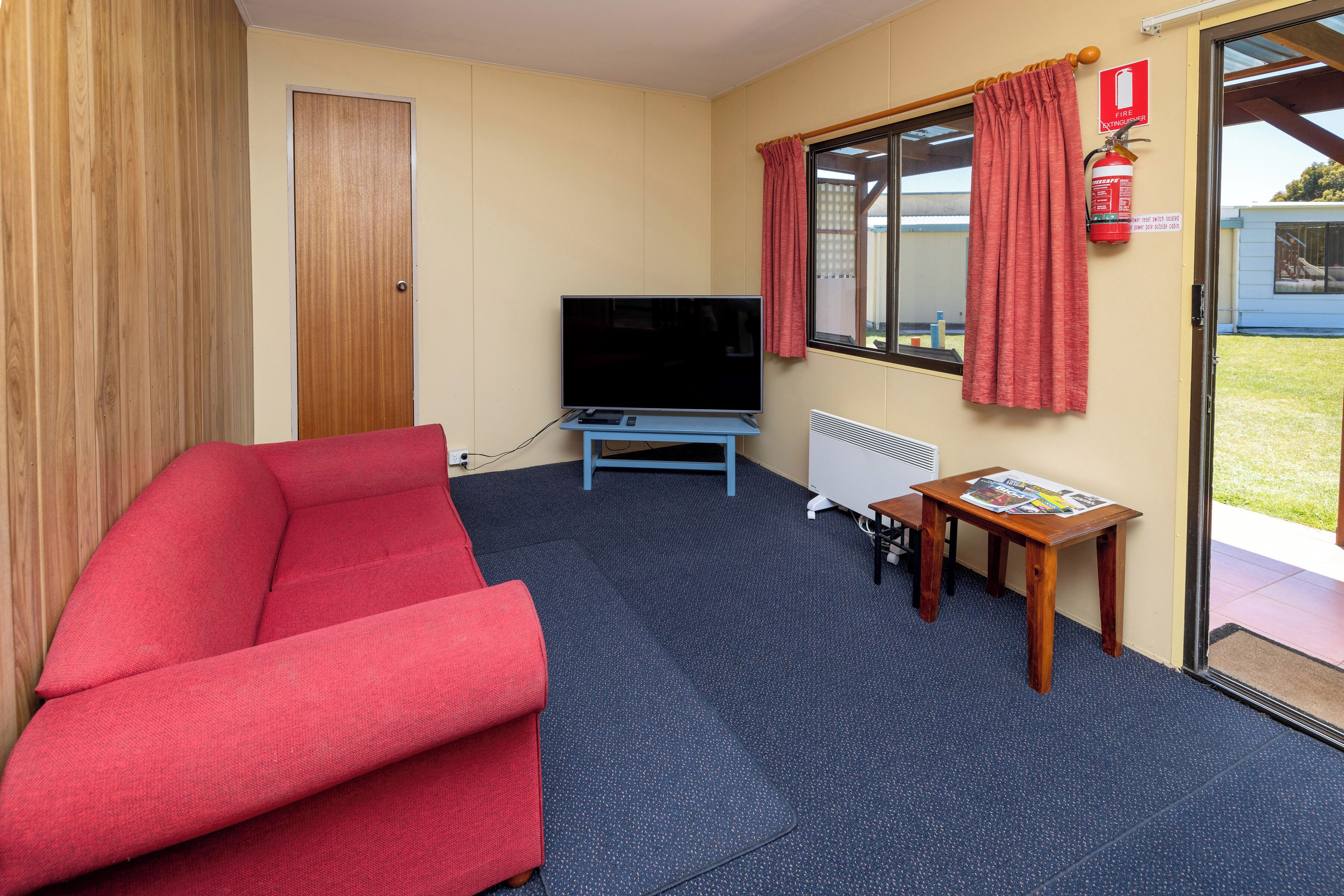 A room with a red sofa, a television, a small wooden table with a newspaper, a radiator, and a window with curtains.