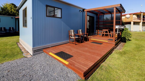 Ein kleines, eingeschossiges Gebäude mit blauer Fassade, einer Holzterrasse und einer überdachten Terrasse mit Gartenmöbeln.