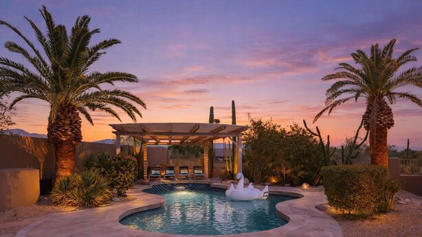 A pool with a swan float, palm trees, a covered seating area, and desert landscaping.