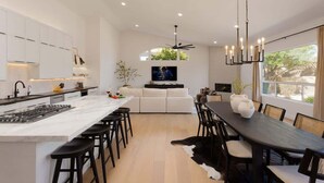 A modern kitchen with a central island, a dining area with a dark wooden table, and a living space with a sofa and TV.