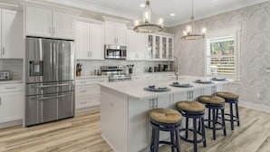 Una cocina moderna con gabinetes blancos, electrodomésticos de acero inoxidable, una isla central con taburetes y una araña.