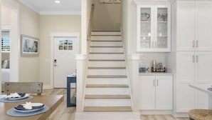 Una escalera conduce a una cocina con gabinetes blancos y un comedor con una mesa y sillas de madera.