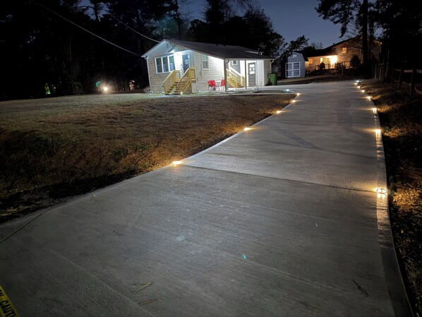 A concrete pathway with string lights leading to a house with a porch and a garage.