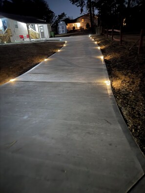 A concrete pathway with evenly spaced lights, flanked by a wooden fence and a small shed.