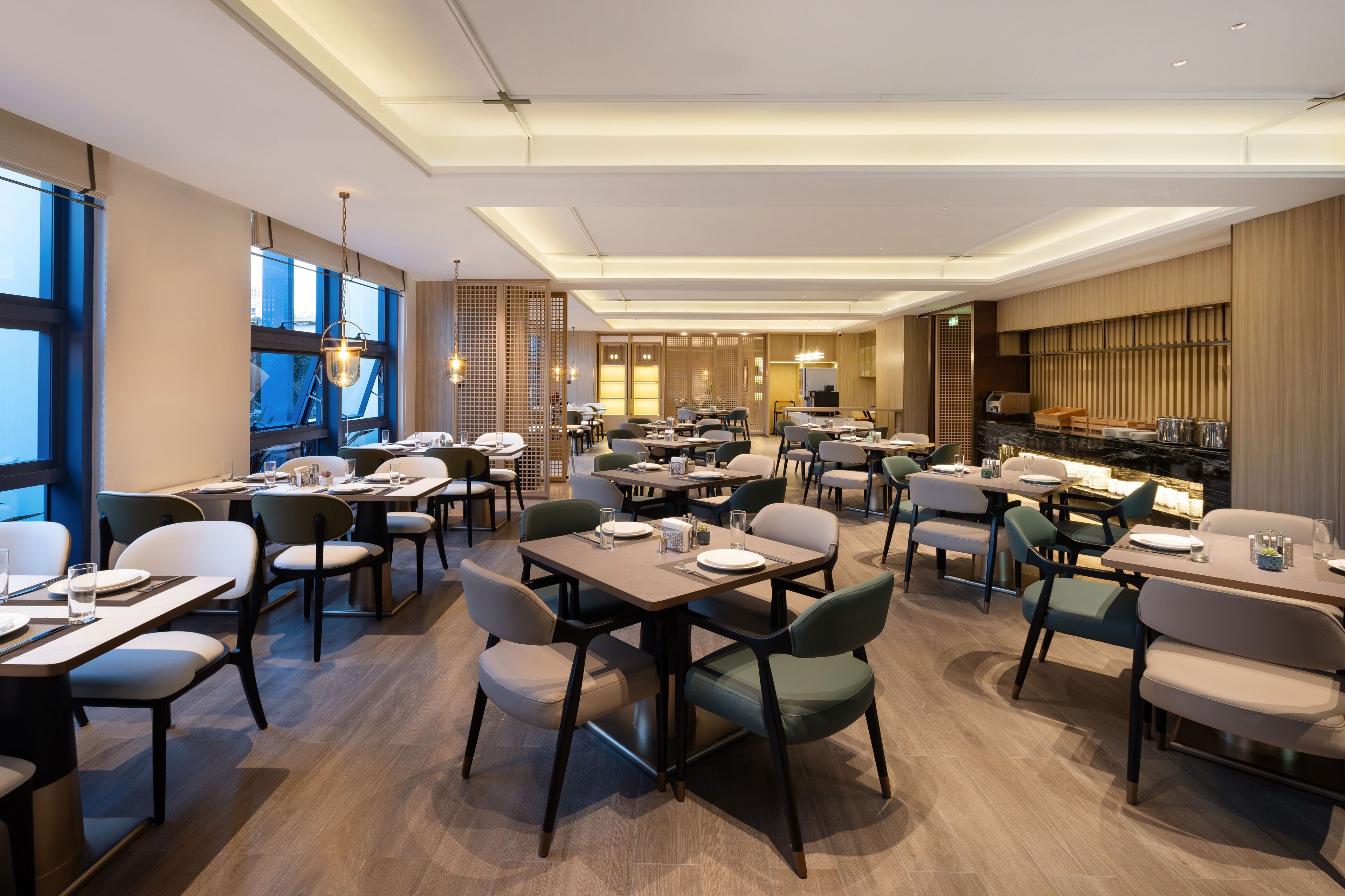Un espace de restauration moderne, doté de plusieurs tables et chaises, de grandes fenêtres et d’une cuisine en arrière-plan.