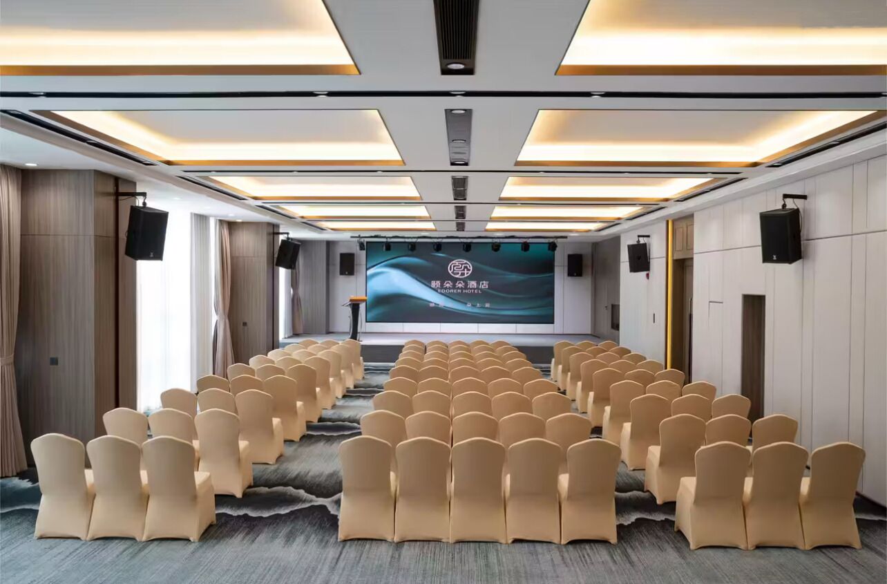 Un salón de conferencias con filas de sillas color beige, una pantalla grande al frente y luces en el techo.