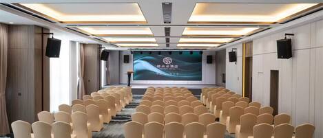 Un salón de conferencias con filas de sillas color beige, una pantalla grande al frente y luces en el techo.
