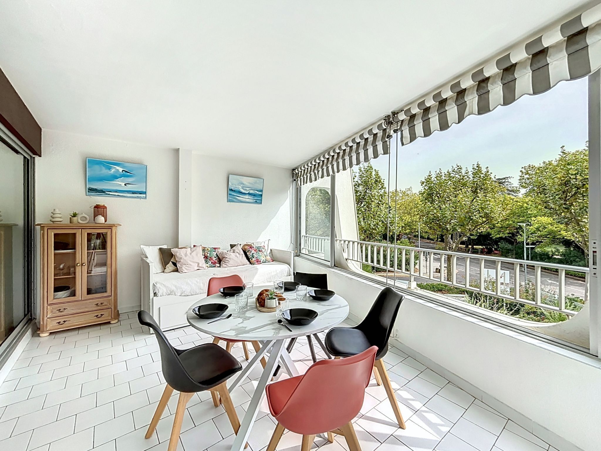 Ein Balkon mit Esstisch, Stühlen und einer Couch. An den Wänden hängen zwei Gemälde und es gibt ein Schrank mit Glastüren.