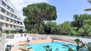 Une piscine avec un grand arbre en arrière-plan, entourée d’une zone délimitée par une clôture, et un bâtiment situé d’un côté.