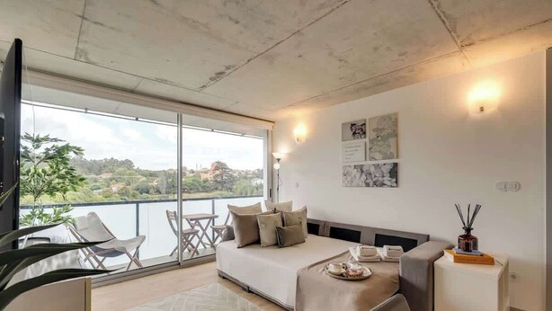 Un dormitorio moderno con una cama grande, un sofá y una mesa de comedor. Cuenta con un balcón con sillas y vistas de la ciudad.