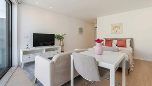 Una habitación moderna, bien iluminada, con televisión de pantalla plana, una mesa de comedor, un sofá y una cama con cabecera.