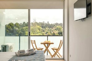 Et soveværelse med balkon, seng med blåt sengetæppe, to træstole, et lille bord med et glas, og et TV ophængt på væggen.