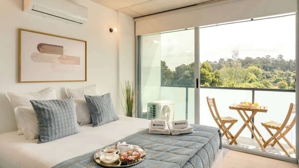 Un dormitorio moderno con una cama grande, vistas a la vegetación a través de un ventanal corredero y una pequeña mesa con sillas afuera.