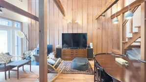 A modern living room with wooden walls, a large flat-screen TV, a dining table, and a cozy seating area with a bench and chairs. - The Cozy Nest MATSUKAWA (Hakodate)