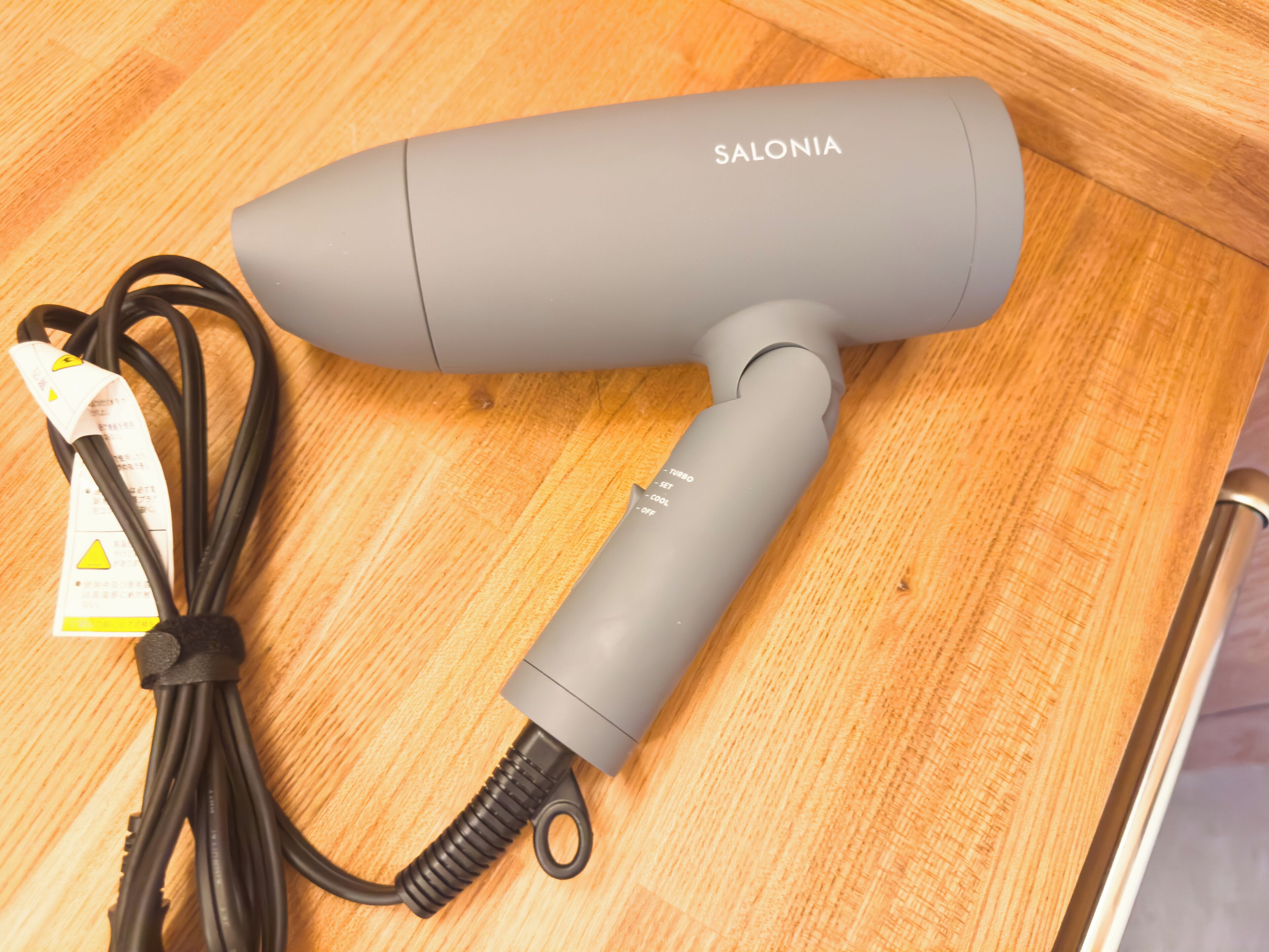 A gray Salonia hairdryer with a power cord and a safety tag on a wooden surface.