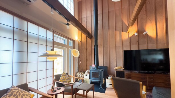 A spacious living room with a high ceiling, wooden walls, a large TV, and a fireplace. - The Cozy Nest MATSUKAWA (Hakodate)