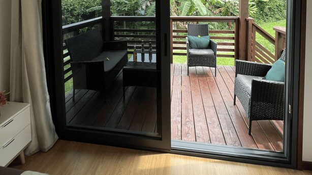 A room with a sliding glass door leading to an outdoor deck with wicker furniture and a view of lush greenery. - Hava'i Lodge (Raiatea)