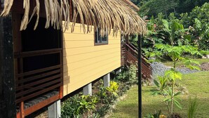 A thatched-roof building with a small window, surrounded by a garden with various plants and a view of a hilly landscape. - Hava'i Lodge (Raiatea)