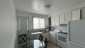 A compact kitchen with white appliances, a glass dining table, and a window providing natural light.