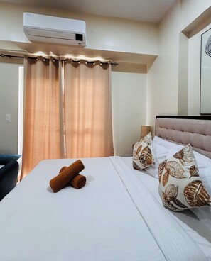 A hotel room with a bed, a window with curtains, an air conditioning unit, and a headboard with patterned pillows. - A Dreamy & Elegant Studio (Iloilo)
