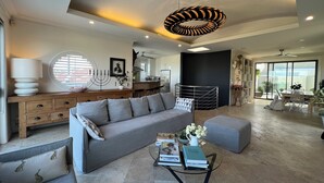 A modern living room with a grey sofa, a coffee table with books, a unique ceiling light, and a view of the kitchen and balcony. - Stunning beach house overlooking North Curl Curl beach (North Curl Curl)