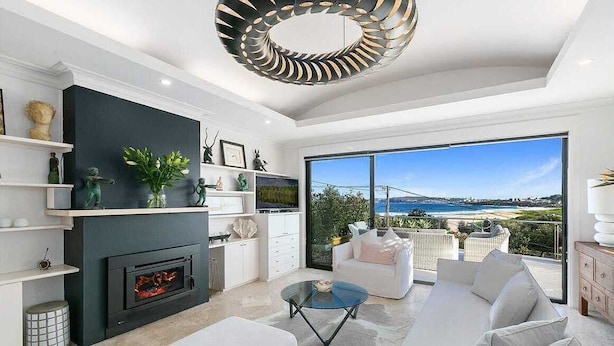 A modern living room with a fireplace, white sofas, a coffee table, and a large window offering a view of the sea. - Stunning beach house overlooking North Curl Curl beach (North Curl Curl)