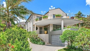 A modern two-story house with a curved balcony, a garage, and lush greenery. - Stunning beach house overlooking North Curl Curl beach (North Curl Curl)
