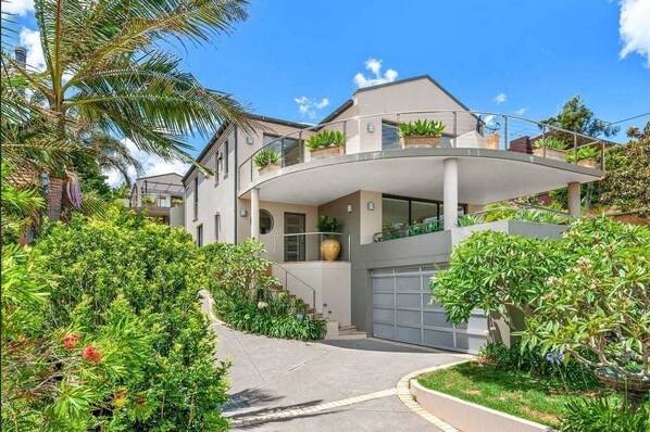 Ein modernes, zweistöckiges Haus mit einem geschwungenen Balkon, einer Garage und üppigem Grün.