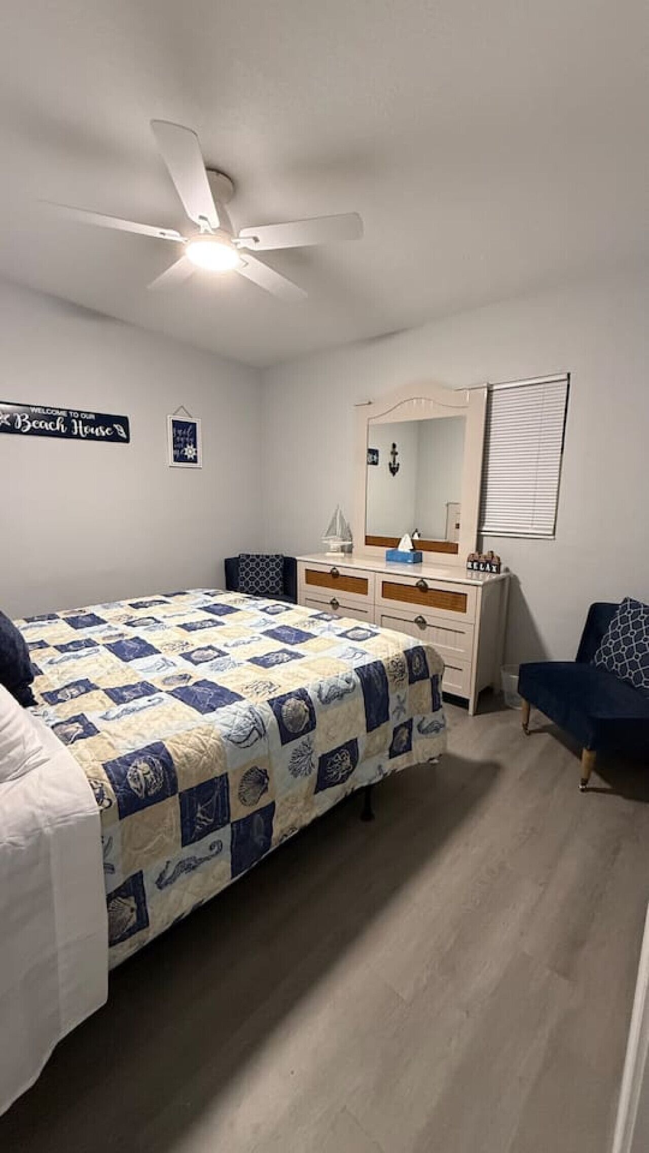 A bedroom with a bed, dresser, and a ceiling fan. There is a sign on the wall that says "Welcome to our Beach House."
