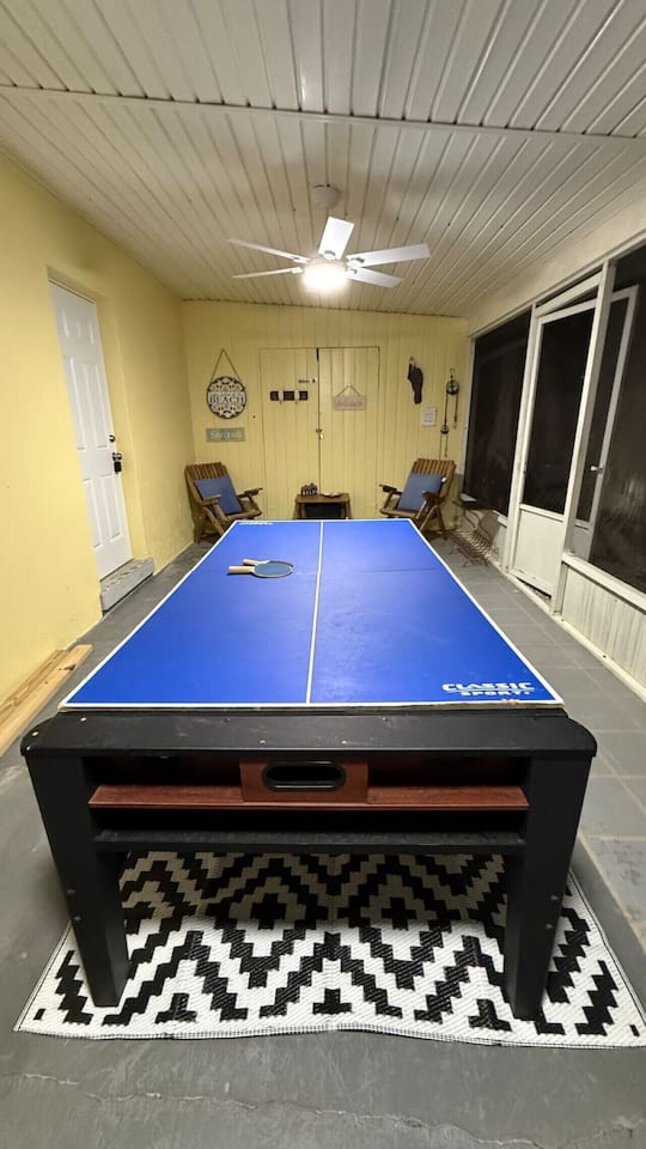 A ping pong table with a black and white patterned rug underneath, a ceiling fan, and a door with a small window.