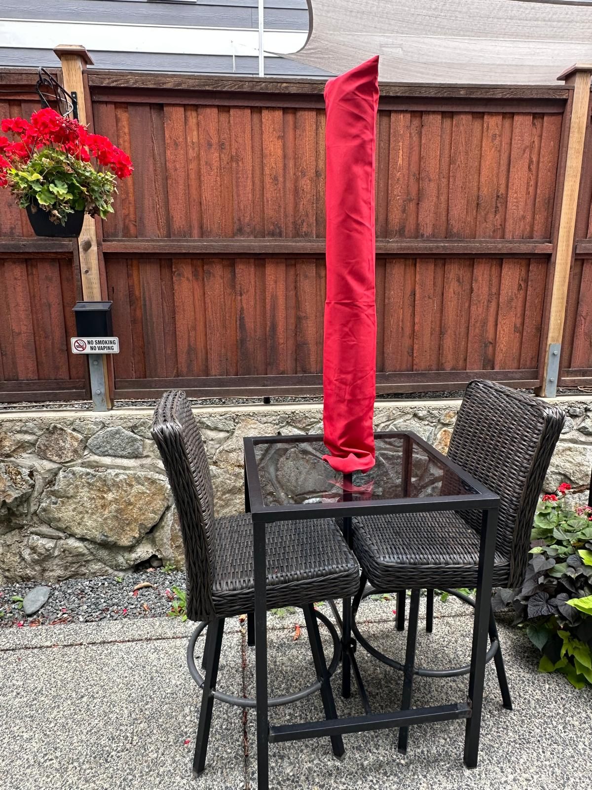 Um pátio com mesa e cadeiras, um guarda-sol vermelho, uma cerca de madeira e uma placa de 'Não Fumar'.