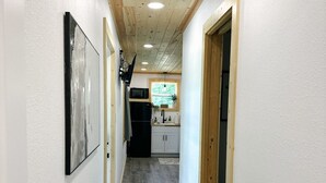 A hallway with a wooden ceiling, white walls, and a rug leading to a kitchen area with cabinets and a window.