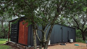 A small, single-story building with a dark exterior, surrounded by trees and a gravel area.