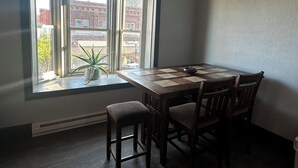 A dining area with a table and chairs, a potted plant on the windowsill, and a view of buildings outside. - Downtown Lofts (International Falls)