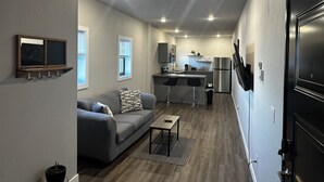 A compact living space with a kitchenette, grey walls, a sofa, and a small table. - Downtown Lofts (International Falls)