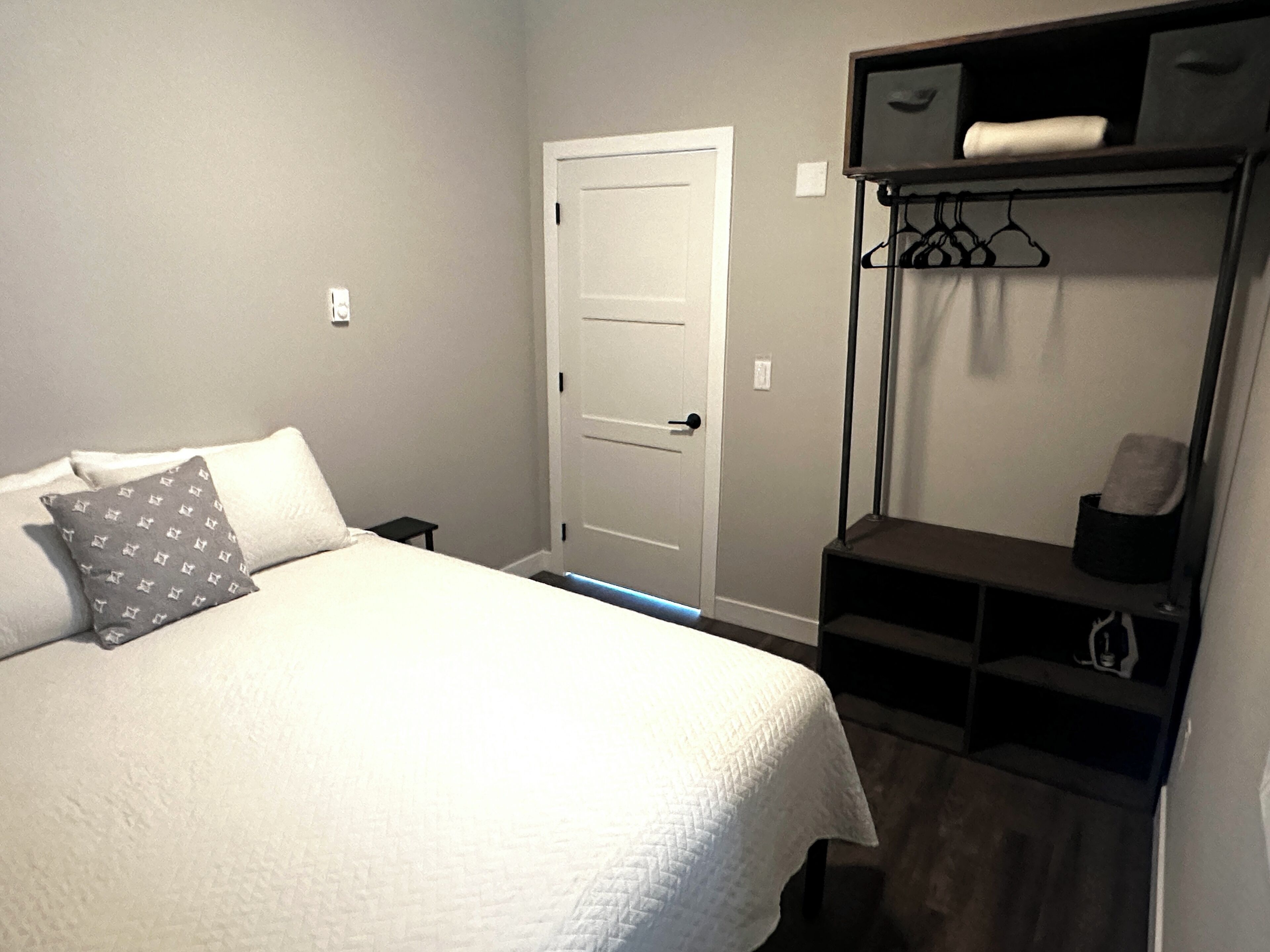 Une chambre à coucher avec un lit, une porte, une étagère où sont suspendus des vêtements et une petite table.