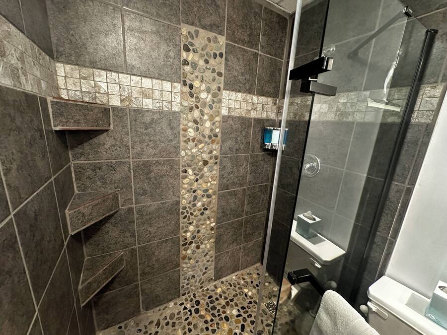 Zona de ducha con puerta de vidrio, suelo de baldosas con diseño de guijarros y mosaico vertical en la pared.