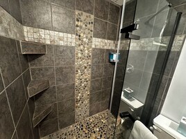 Zona de ducha con puerta de vidrio, suelo de baldosas con diseño de guijarros y mosaico vertical en la pared.