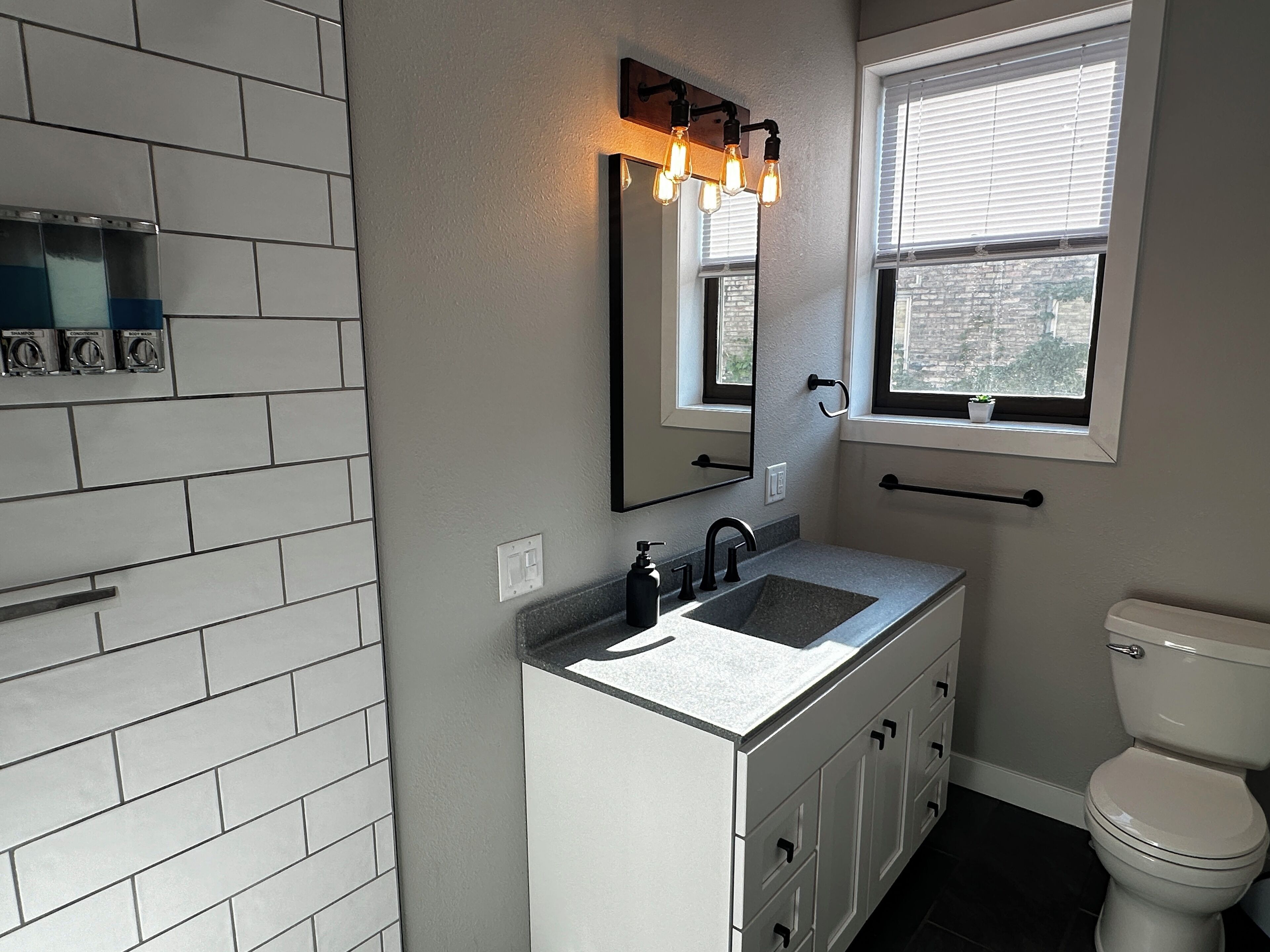 Een badkamer met een witte wastafel, een zwarte wasbak, een spiegel met verlichting, een raam met jaloezieën en een toilet.
