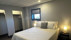 A neatly made bed with a quilted cover, two bedside lamps, a window with blinds, and a coat rack. - Downtown Lofts (International Falls)