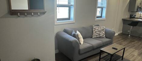 A modern living room with a grey sofa, a small coffee table, and a kitchen area with a bar and a microwave.