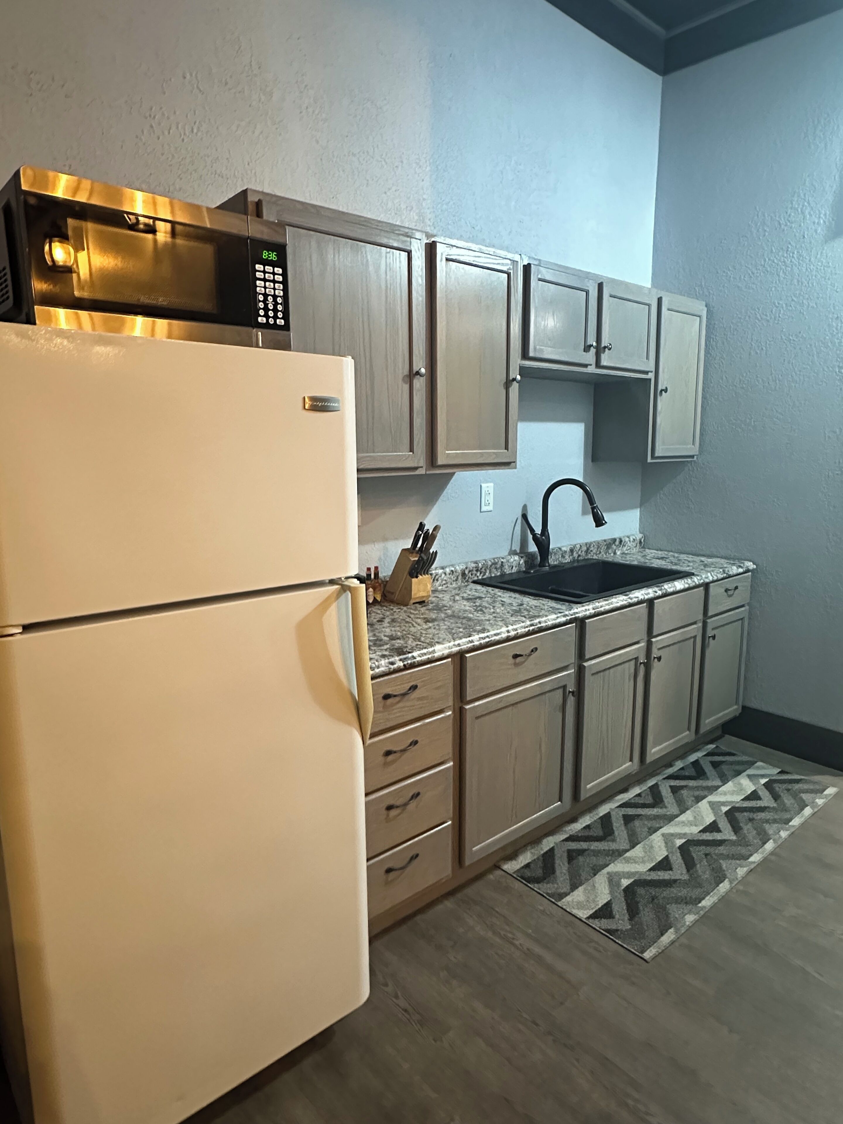 A kitchen with a white refrigerator, a microwave, wooden cabinets, a granite countertop, a sink, and a patterned rug.