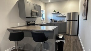 A modern kitchen with a central island, two bar stools, stainless steel appliances, and a trash can. - Downtown Lofts (International Falls)