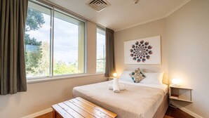 A hotel room with a large bed, a wooden bench, a nightstand with a lamp, and a view of trees through the window. - Auchenflower 1BR Retreat, Parking Close to UQ & M5 (Auchenflower)