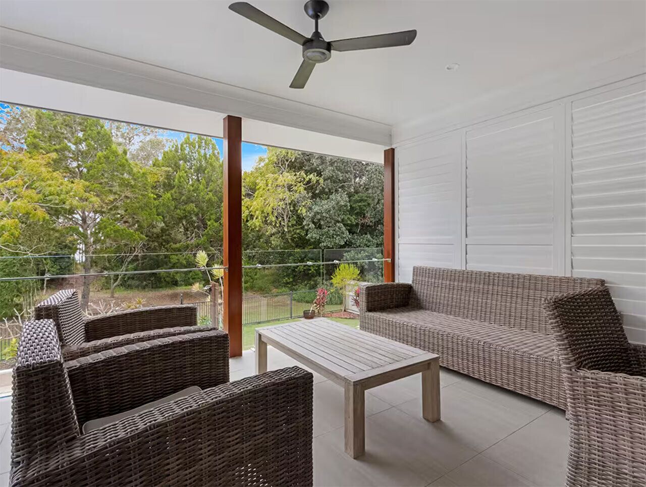 A covered outdoor seating area with wicker furniture, a ceiling fan, and a view of the surrounding greenery.